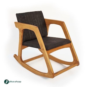 Peut inclure: Une chaise &agrave; bascule dot&eacute;e d'une structure en bois brun clair avec une finition grain naturel et un si&egrave;ge ainsi qu'un dossier rembourr&eacute;s gris fonc&eacute;. Les coussins pr&eacute;sentent des lignes cousues verticales et la marque Pikus Ahşap est visible.