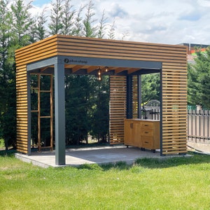 May include: A wooden pergola with a slatted roof and black metal supports. The pergola has a wooden cabinet with drawers underneath. The text "pikkus ahşap" is visible on the pergola.