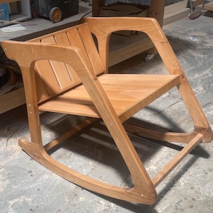 Peut inclure: Une chaise &agrave; bascule en bois clair dot&eacute;e d'un dossier &agrave; lattes et d'une assise plate. La chaise est construite avec des supports lat&eacute;raux inclin&eacute;s reli&eacute;s &agrave; des patins incurv&eacute;s &agrave; la base, avec des fixations m&eacute;talliques visibles aux jointures.