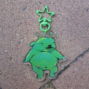 Può includere: Un portachiavi in acrilico verde lime raffigurante una creatura aliena dei cartoni animati con un corpo rotondo, due antenne e un'espressione con gli occhi chiusi. Il ciondolo è fissato a un moschettone girevole in plastica a stella.