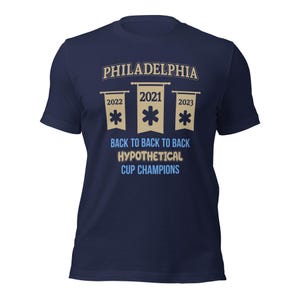 Puede incluir: Una camiseta azul marino con un gráfico de tres pancartas. Las pancartas son beige con texto azul y una estrella azul. El texto en las pancartas dice "2021", "2022" y "2023". El texto debajo de las pancartas dice "Philadelphia, Back to Back to Back Hypothetical Cup Champions".