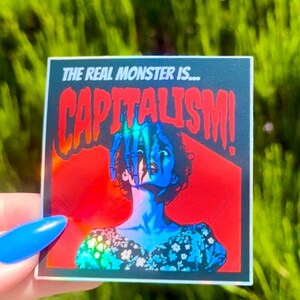 Puede incluir: Una pegatina hologr&aacute;fica con fondo rojo y negro que representa a una mujer con una mano que sale de detr&aacute;s de ella. El texto de la pegatina dice "The real monster is... CAPITALISM!"