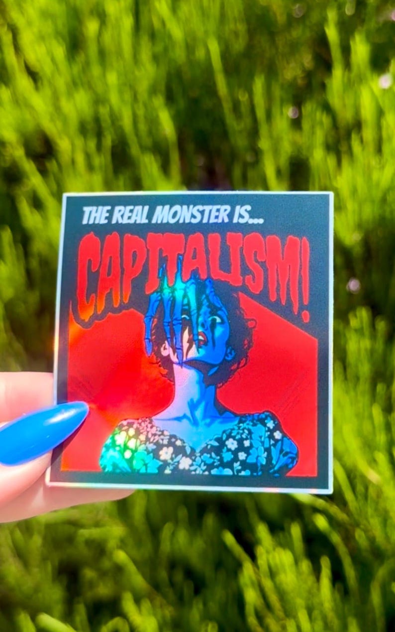 Puede incluir: Una pegatina hologr&aacute;fica con fondo rojo y negro que representa a una mujer con una mano que sale de detr&aacute;s de ella. El texto de la pegatina dice "The real monster is... CAPITALISM!"