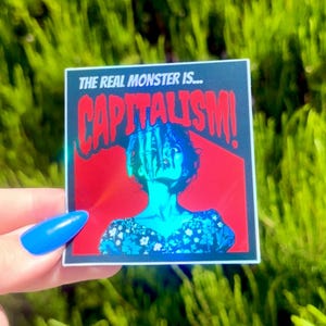 Puede incluir: Una pegatina hologr&aacute;fica con fondo rojo y borde negro. La pegatina muestra a una mujer con una mano que le llega a la cara. El texto de la pegatina dice "The real monster is... CAPITALISM!"