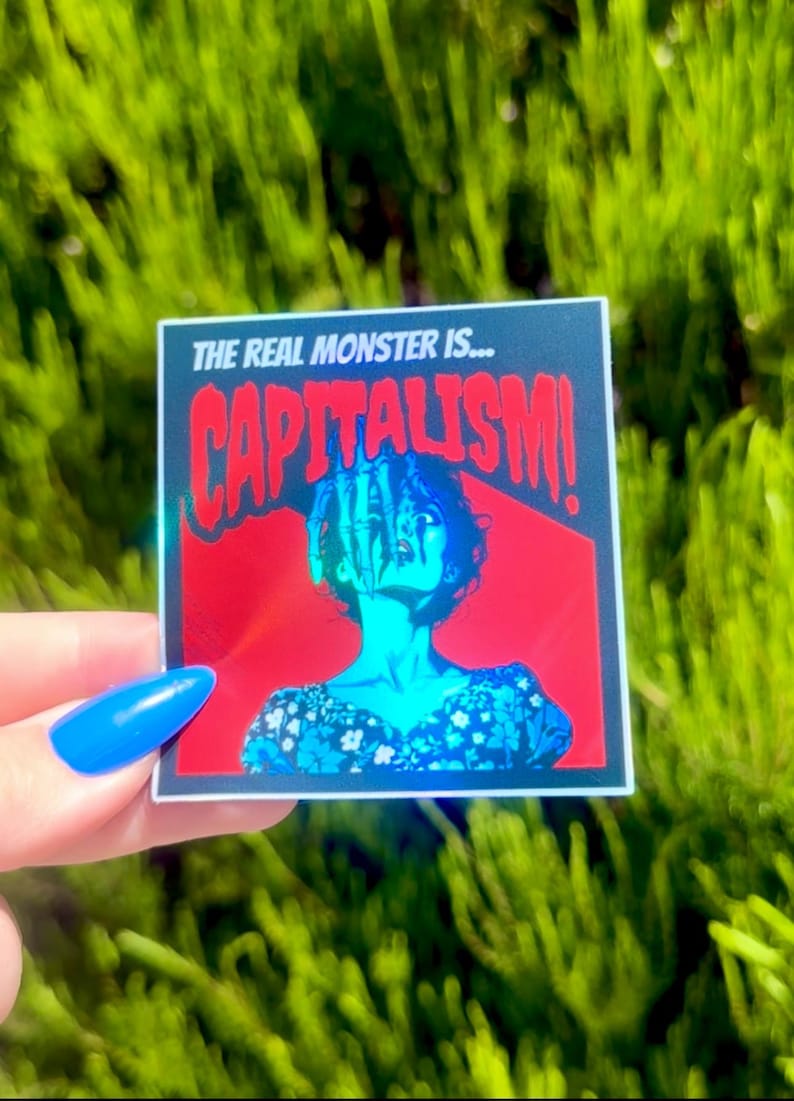 Puede incluir: Una pegatina hologr&aacute;fica con fondo rojo y borde negro. La pegatina muestra a una mujer con una mano que le llega a la cara. El texto de la pegatina dice "The real monster is... CAPITALISM!"