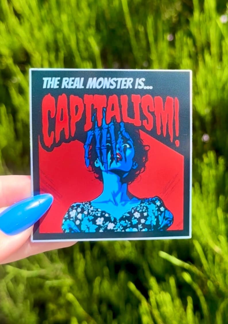 Puede incluir: Una pegatina con fondo rojo y negro que muestra a una mujer con una mano que le llega a la cara. El texto "THE REAL MONSTER IS... CAPITALISM!" est&aacute; impreso en blanco.