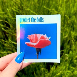 May include: A square sticker with a holographic border featuring a coral rose against a bright blue background. The text "protect the dolls" is printed in light blue at the top. The sticker is held against a backdrop of green foliage.
