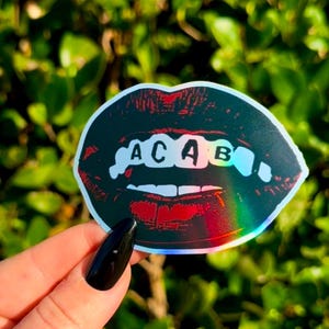 May include: A black and red holographic sticker of a pair of lips with the word "ACAB" spelled out in white letters on the teeth.
