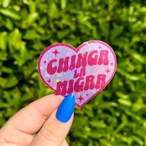 Può includere: Un adesivo a forma di cuore con la scritta "CHINGA LA MIGRA" in lettere rosa glitterate. L'adesivo ha uno sfondo olografico con accenti di stelle. Lo sfondo è verde sfocato.