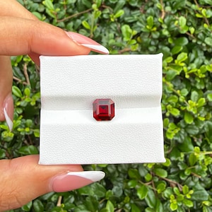 May include: A deep red, emerald-cut gemstone with sharp, faceted edges, securely nestled in the center of a white, rectangular display box featuring a horizontal slit across its textured surface for holding the stone in place.