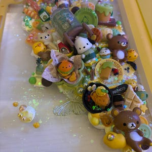 May include: A yellow phone case decorated with miniature food items, bears, and other cute figurines. The case features a variety of colors, including brown, yellow, green, and white, with glitter accents. The case is filled with small, detailed objects.