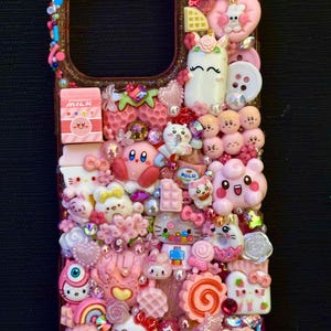 Puede incluir: Una funda de teléfono rosa adornada con una variedad de elementos decorativos. La funda presenta personajes de dibujos animados, fresas, corazones y un macaron. La funda está adornada con pedrería y otros pequeños detalles coloridos.