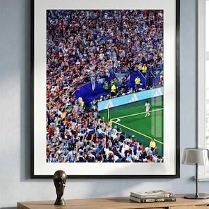 May include: A framed photograph of a soccer match featuring a player in a white jersey preparing to take a corner kick on a green field. A large crowd of spectators in blue and white clothing fills the stands, and a gold trophy replica sits nearby.