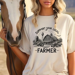 Puede incluir: Una mujer lleva una camiseta beige claro con un gráfico negro de una escena de granja y el texto "Support Your Local Farmer". La mujer está sosteniendo un caballo marrón y blanco con una cabezada negra. La mujer lleva un sombrero beige claro. El fondo es una imagen borrosa de un campo y una colina.