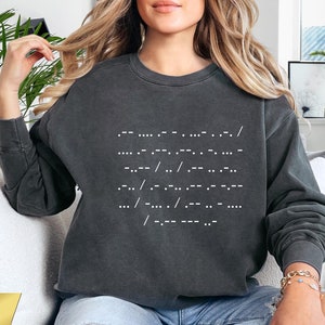 Może przedstawiać: Szara bluza z ciemnym odcieniem z białym nadrukiem frazy "Whatever happens, I will always be with you" w alfabecie Braille'a. Bluzę nosi osoba siedząca na białej kanapie.