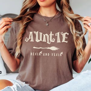 Pode incluir: Camiseta marrom com a palavra "Auntie" em uma fonte decorativa, um gráfico de vassoura e a frase "Hexin and Yexin". A camiseta tem um ajuste confortável e é perfeita para o Halloween ou um evento temático de bruxas.