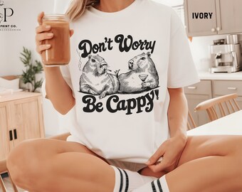 May include: White t-shirt with a graphic of two capybaras, one holding a glass of wine and the other smoking a cigarette. The text "Don't Worry Be Cappy!" is printed in black below the image. The shirt is on a person sitting on a white table.