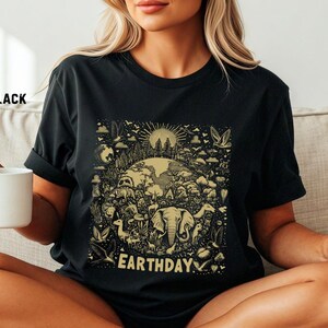 Könnte beinhalten: Schwarzes T-Shirt mit einem Earth Day-Design. Die detaillierte Grafik zeigt die Erde, Tiere, Bäume und den Text "EARTHDAY". Das T-Shirt besteht aus einem weichen Material.