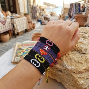 May include: A collection of colorful woven bracelets featuring superhero logos. The bracelets are black, purple, and red, with white, yellow, and red embroidered designs. The bracelets are stacked on a wrist.