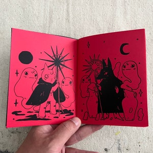 Puede incluir: Dos p&aacute;ginas abiertas de un peque&ntilde;o libro rojo con ilustraciones en tinta negra. La p&aacute;gina izquierda muestra una criatura parecida a un gato que lleva una capa y sostiene un bast&oacute;n en forma de estrella. La p&aacute;gina derecha muestra una criatura parecida a un lobo que lleva una capa y sostiene un bast&oacute;n en forma de estrella. Ambas p&aacute;ginas presentan fantasmas y otros elementos m&iacute;sticos.