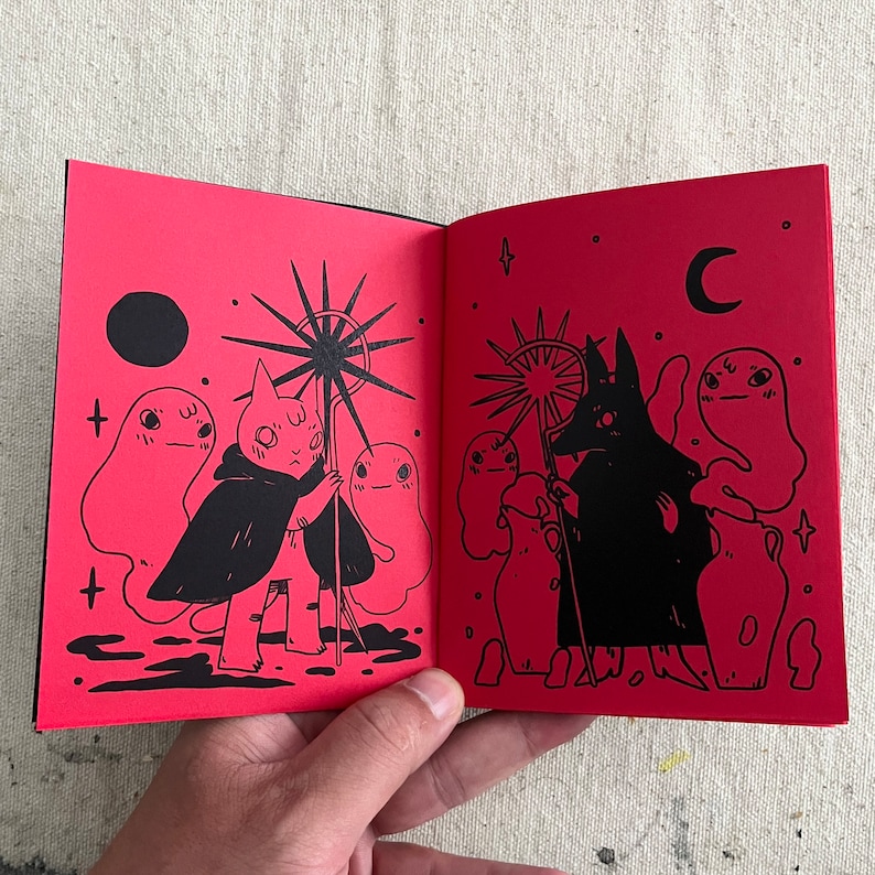 Puede incluir: Dos p&aacute;ginas abiertas de un peque&ntilde;o libro rojo con ilustraciones en tinta negra. La p&aacute;gina izquierda muestra una criatura parecida a un gato que lleva una capa y sostiene un bast&oacute;n en forma de estrella. La p&aacute;gina derecha muestra una criatura parecida a un lobo que lleva una capa y sostiene un bast&oacute;n en forma de estrella. Ambas p&aacute;ginas presentan fantasmas y otros elementos m&iacute;sticos.