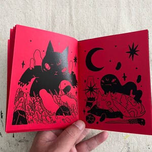 Puede incluir: Un libro rojo con ilustraciones negras de gatos y otras criaturas. El libro est&aacute; abierto en una p&aacute;gina con un gato negro grande con una luna creciente y estrellas en el fondo.