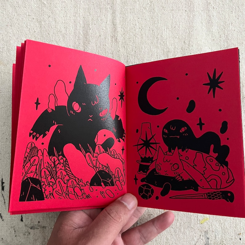 Puede incluir: Un libro rojo con ilustraciones negras de gatos y otras criaturas. El libro est&aacute; abierto en una p&aacute;gina con un gato negro grande con una luna creciente y estrellas en el fondo.