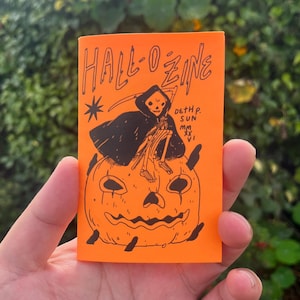 May include: A bright orange zine titled HALL-O-ZINE in black ink. The cover features a black illustration of a hooded skeleton figure holding a scythe while sitting atop a carved jack-o'-lantern pumpkin. Text reads DETH P. SUN MMXXVI.