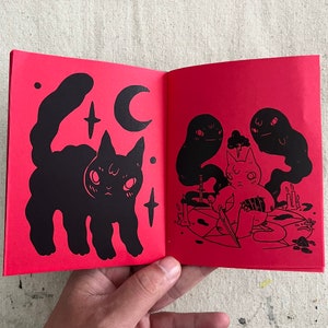 Puede incluir: Un libro ilustrado rojo con un gato negro en la p&aacute;gina izquierda y un gato negro con una espada en la p&aacute;gina derecha. El gato de la p&aacute;gina derecha est&aacute; rodeado de fantasmas.