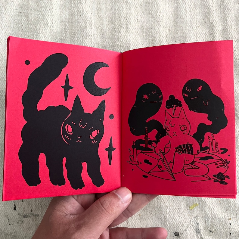 Puede incluir: Un libro ilustrado rojo con un gato negro en la p&aacute;gina izquierda y un gato negro con una espada en la p&aacute;gina derecha. El gato de la p&aacute;gina derecha est&aacute; rodeado de fantasmas.