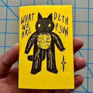 May include: A rectangular yellow card featuring a black cat illustration with a visible yellow skeletal and organ structure. The text WHAT WE ARE DETH P.SUN is printed in black ink. A small four-pointed star symbol is located to the right of the cat.