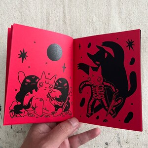 Puede incluir: Un libro abierto con ilustraciones negras de gatos sobre un fondo rojo. La p&aacute;gina izquierda muestra tres gatos, uno con un esqueleto, y la p&aacute;gina derecha muestra un gato con un esqueleto y la sombra de un gato.