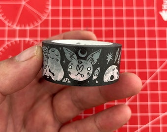 May include: Black and white washi tape with a repeating pattern of a bat, moon, stars, and ghosts. The tape is rolled up and held in a hand.