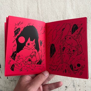 Puede incluir: Un libro abierto con p&aacute;ginas rojas e ilustraciones negras. La p&aacute;gina izquierda muestra un gato con una tetera, y la p&aacute;gina derecha muestra una espada, cr&aacute;neos y una criatura.