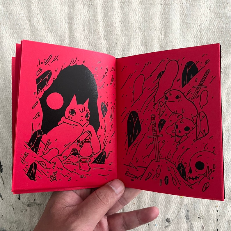 Puede incluir: Un libro abierto con p&aacute;ginas rojas e ilustraciones negras. La p&aacute;gina izquierda muestra un gato con una tetera, y la p&aacute;gina derecha muestra una espada, cr&aacute;neos y una criatura.