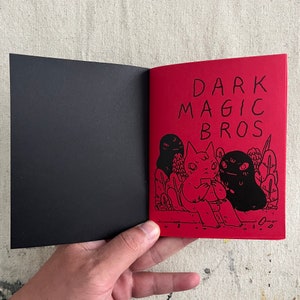 Puede incluir: Un c&oacute;mic rojo y negro con el t&iacute;tulo "DARK MAGIC BROS" en la portada. La portada presenta una ilustraci&oacute;n en blanco y negro de tres personajes de dibujos animados, dos de los cuales visten ropa blanca y negra y uno de los cuales viste ropa roja y blanca.