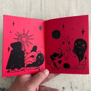 Puede incluir: Dos p&aacute;ginas de un libro ilustrado en blanco y negro con fondo rojo. La p&aacute;gina izquierda muestra una figura encapuchada con un bast&oacute;n de pie frente a una explosi&oacute;n de estrellas. La p&aacute;gina derecha muestra un gato sentado en un cr&aacute;neo con un fantasma detr&aacute;s.