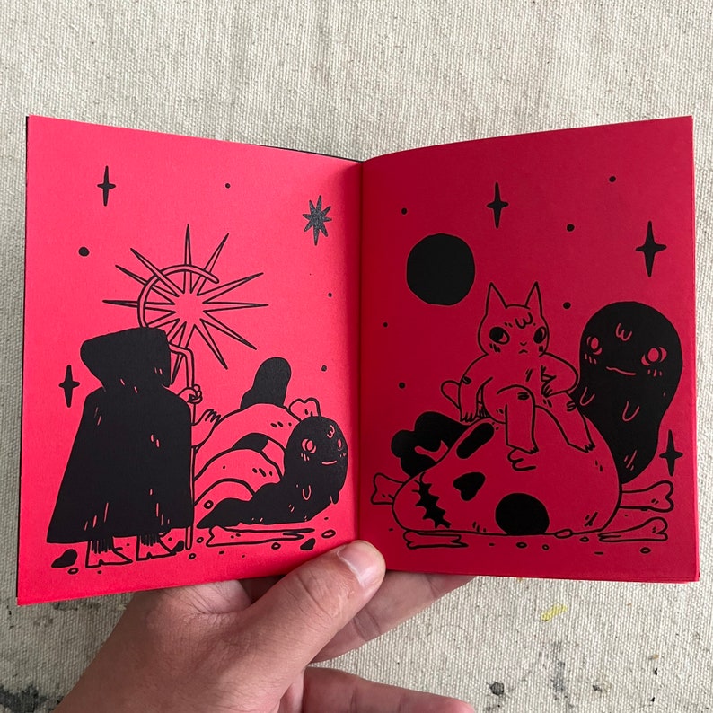 Puede incluir: Dos p&aacute;ginas de un libro ilustrado en blanco y negro con fondo rojo. La p&aacute;gina izquierda muestra una figura encapuchada con un bast&oacute;n de pie frente a una explosi&oacute;n de estrellas. La p&aacute;gina derecha muestra un gato sentado en un cr&aacute;neo con un fantasma detr&aacute;s.