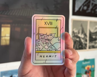 May include: A rectangular sticker featuring a tarot card design. The card is labeled "HERMIT" and "XVIII" in black text. The illustration inside the card depicts a group of creatures. The sticker has a holographic, iridescent finish with pastel colors.