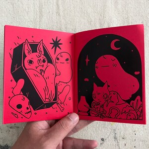 Puede incluir: Un libro abierto con p&aacute;ginas rojas e ilustraciones negras. La p&aacute;gina izquierda muestra un murci&eacute;lago en un ata&uacute;d con un fantasma. La p&aacute;gina derecha muestra un fantasma con un gato y un p&aacute;jaro.