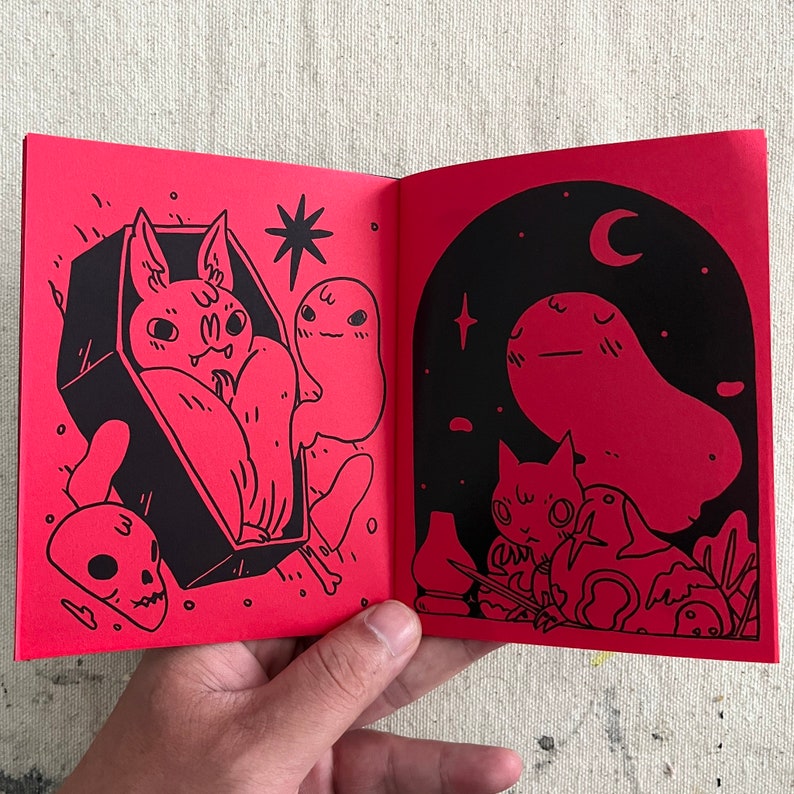 Puede incluir: Un libro abierto con p&aacute;ginas rojas e ilustraciones negras. La p&aacute;gina izquierda muestra un murci&eacute;lago en un ata&uacute;d con un fantasma. La p&aacute;gina derecha muestra un fantasma con un gato y un p&aacute;jaro.