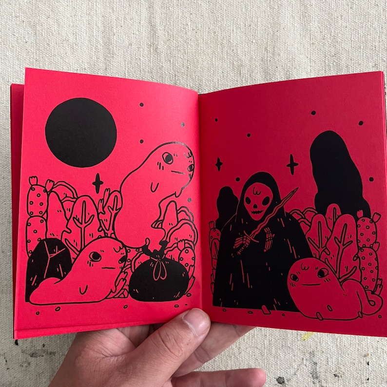 Puede incluir: Un libro ilustrado rojo con dibujos en tinta negra de una figura de esqueleto, un fantasma y otras criaturas. El libro est&aacute; abierto en una p&aacute;gina con fondo negro y estrellas blancas.