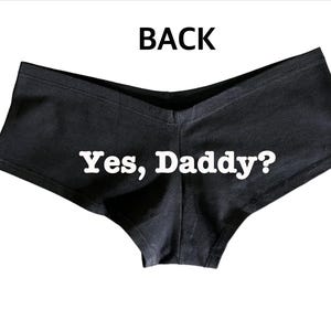 Pode incluir: Roupa interior preta com o texto "Yes, Daddy?" em branco. A roupa interior é de estilo de cintura baixa com uma cintura em forma de V. A palavra "BACK" está impressa na parte superior da imagem.