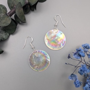 Może przedstawiać: Para srebrnych kolczyków z iryzującymi, okrągłymi zawieszkami w kształcie muszli. Kolczyki mają prosty, srebrny wzór haka.