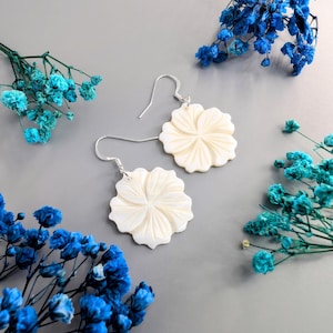 May include: Cream-coloured flower-shaped earrings with silver hooks. The earrings are set against a grey background, with blue and turquoise dried flowers. The earrings have a detailed, carved petal design.