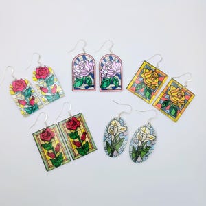 Puede incluir: Una colección de pendientes coloridos estilo vidriera con diseños florales. Los pendientes muestran rosas y calas en varias formas, incluyendo arqueadas, rectangulares y ovaladas. Cada pendiente tiene un gancho plateado.