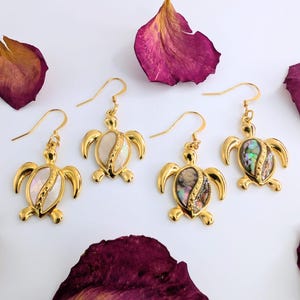 May include: Four gold-coloured turtle earrings with iridescent shells and gold hooks. The earrings are displayed on a white surface with scattered dried, dark pink rose petals. The turtle design features a shell-shaped inlay.
