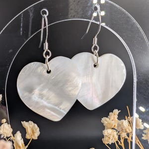 May include: A pair of heart-shaped earrings with a pearlescent, iridescent finish. The earrings are suspended from silver-colored fish hook ear wires. The earrings are displayed against a black background with dried flowers.