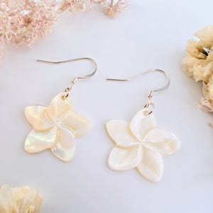 Könnte beinhalten: Ein Paar blütenförmige Ohrringe mit Perlmutt-Finish. Die Ohrringe sind weiß mit einem dezenten schillernden Glanz und hängen an silberfarbenen Haken. Das florale Design hat fünf Blütenblätter.