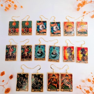 Puede incluir: Un conjunto de pendientes colgantes dorados con diseños de cartas del tarot. Cada pendiente muestra una carta diferente, incluyendo "El Sol", "La Luna", "La Emperatriz" y "El Loco". Las cartas tienen ilustraciones coloridas y ganchos dorados.